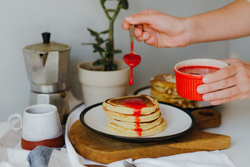 Girl pouring raspberry sauce on pancakes near coffee maker on white table in kitchen. Morning breakfast at home in quarantine. Recipe of homemade delicious pancakes.
