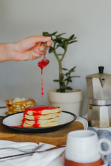 Girl pouring raspberry sauce on pancakes near coffee maker on white table in kitchen. Morning breakfast at home in quarantine. Recipe of homemade delicious pancakes.