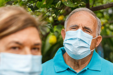 Anziano con mascherina chirurgica guarda serio in macchina, in primo piano la moglie sfocata. Isolati su sfondo natura