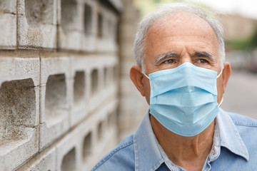 Anziano con mascherina chirurgica guarda serio vicino alla recinzione di una casa