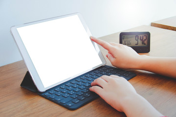 Image of hands taping on a tablet with white blank screen which has a keyboard attach to it. Wood table with clock and white wall.