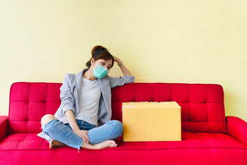 Young asian woman wearing mask sitting on red couch at home with feeling sad and disheartened from company layoff during covid-19, reduction cost. Pandemic disease and impacts every business.