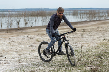 Fototapeta premium Sports brutal bearded guy on a modern mountain bike. A cyclist in a salt deserted place by the lake.