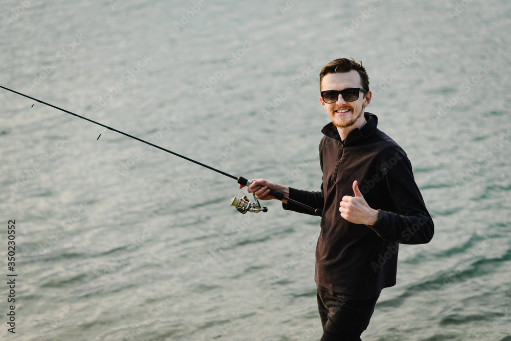 Wall mural Fishing for pike, perch, carp. Fisherman with rod, spinning reel on river bank. Man catching fish, pulling rod while fishing on lake, pond with text space. Wild nature. The concept of rural getaway.