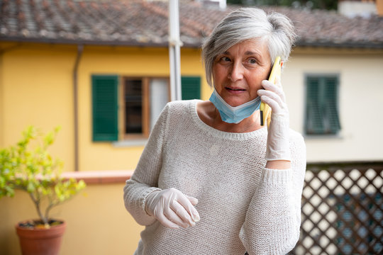 Concept of protection and prevention from Covid-19 Coronavirus - Friendly mature woman call the family with the smartphone wearing latex gloves, medical mask against diffusion of Corona virus