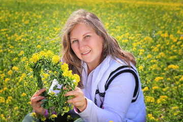 Naklejka na ściany i meble A woman in a white blouse with a yellow flower wreath in her hands is sitting in a flower field. Outdoor recreation. The woman is happy, smiling, happy. Spring, love, joy. The quarantine is over, peop