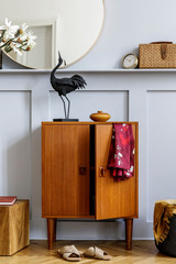 Modern concept of home staging with design wooden commode, round mirror, shelf, clock, plant, cube, clothes, books, rattan bag and elegant personal accessories at stylish living room.