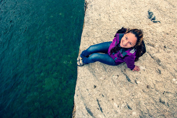 A smiling girl sits on the edge of a stone near the water. European appearance. Jacket and jeans. The water is turquoise clear. Copy space. Horizontal.