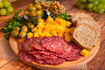 Close-up on a wooden Board, olives, herbs, walnuts, sliced smoked sausage, cheese, bread, green grapes and lemon in the background.