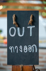 A pavement sign that says &lsquo;OUT&rsquo; and Thai languages on cafe or restaurant hang on a wooden post at entrance.
