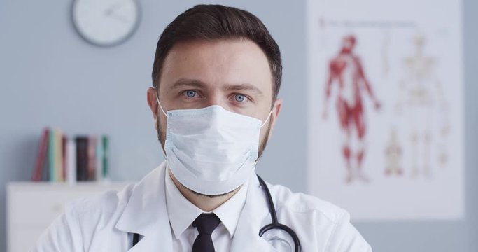 Close-up portrait of of attractive young man doctor in medical mask and statoscope in hospital office looking at camera with serious face. Medics, infection and protection concept.
