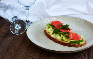Large toast of rye bread with cheese, cabbage, parsley, fresh cucumber and red fish salmon on wooden background with white cloth drapery.