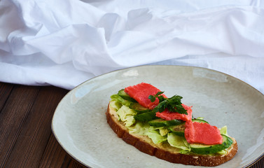 Large toast of rye bread with cheese, cabbage, parsley, fresh cucumber and red fish salmon on wooden background with white cloth drapery.