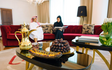 Arab family sitting on a background of dates, raisins and tea waiting for Iftar. 
A man in white traditional clothes and a woman in black abaya chatting at home sitting on the sofa 