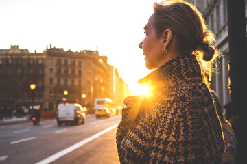 An adult beautiful lady in a plaid coat in the rays of the setting sun stands along the street of barcelona and looks away. View assembly close.