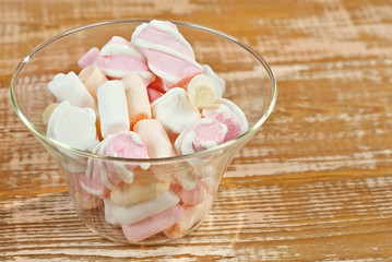 Sweets in a glass on a wooden table. Transparent utensils with marshmallows and biz on a shabby old board. Pink with white soft candies close up.