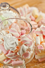 Sweets in a glass on a wooden table. Transparent utensils with marshmallows and biz on a shabby old board. Pink with white soft candies close up.
