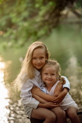 Beautiful girl of 7 years old with long blonde hair with a little sister walks and cuddles at sunset in a park near a pond on a summer evening