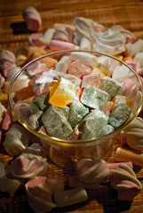 Sweets in a glass on a wooden table. Transparent utensils with marshmallows and biz on a shabby old board. Pink with white soft candies close up.