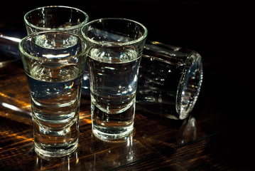Glasses with alcohol near the bottle. Transparent drink on a brown wooden background. Table made of dark shabby boards. Blue reflexes and bright reflections of light on the bottle.