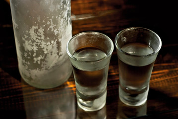 Frozen glasses with alcohol near the bottle. Drinks on a dark brown table. Transparent utensils on a black background. Old shabby boards under glass.