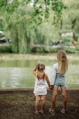 Fototapeta na wymiar Beautiful girl of 7 years old with long blonde hair with a little sister walks and cuddles at sunset in a park near a pond on a summer evening
