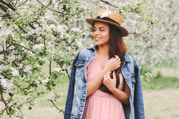 Portrait young cute hippie girl in stylish denim jacket and straw hat relaxing on spring countryside garden with blossom trees. Beautiful happy hipster woman posing on green meadow. Enjoy nature