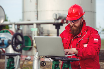 Engineer wearing safety helmet using laptop with oil refinery industry plant background.