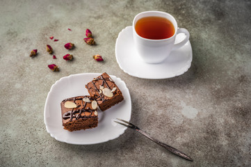 Homemade brownie with almonds on a saucer with a fork on wooden table. Next cup of tea and rose buds. Copy space for text