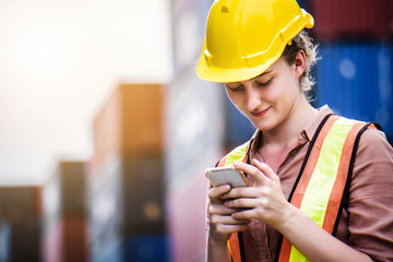 Young confident Caucasian woman engineer using mobile phone and wearing yellow safety helmet and check for control loading containers box from Cargo freight ship for import and export