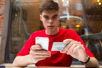 Young man pays for purchase online using cell telephone and credit debit card while sitting in coffee shop during recreation time in weekend