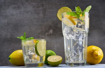 Cold refreshing limonade, mojito or gin tonic in glass, with fresh mint and ice cubes, lime and lemon on wooden board and black background. Summer cocktails and drinks concepts. Copy space for text.