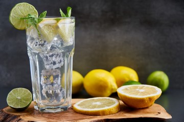 Cold refreshing limonade, mojito or gin tonic in glass, with fresh mint and ice cubes, lime and lemon on wooden board and black background. Summer cocktails and drinks concepts. Copy space for text.