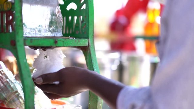 side view of indian ice gola maker manually grinding ice into his hands using an ice grinding machine with bottles filled with colorful syrup in the background.