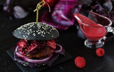 Black burger with raspberry homemade vinaigrette. On a dark background. Inside the cutlet, red onions, purple basil, red cabbage and raspberry homemade vinaigrette. An unusual combination.
