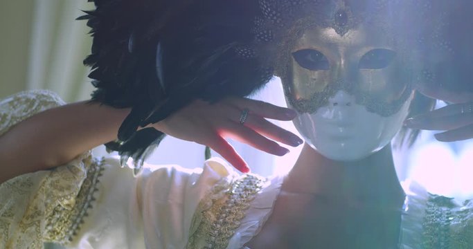 Girl in a golden Venetian mask with black feathers and a vintage dress elegantly moves her hands. The sun shines from the window and the sun's rays fall into the lens