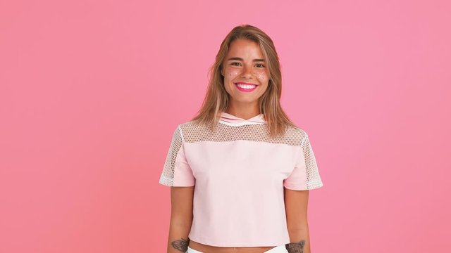 Blonde lady with shiny freckles, dressed in top. She threw back her hair, smiling, showing her palms, posing against pink background. Close up
