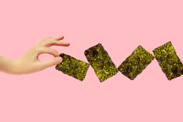Female hand hold crispy seaweed on pink background with copy space. Dry nori sheets korean or japanese roasted food texture. Flat lay.