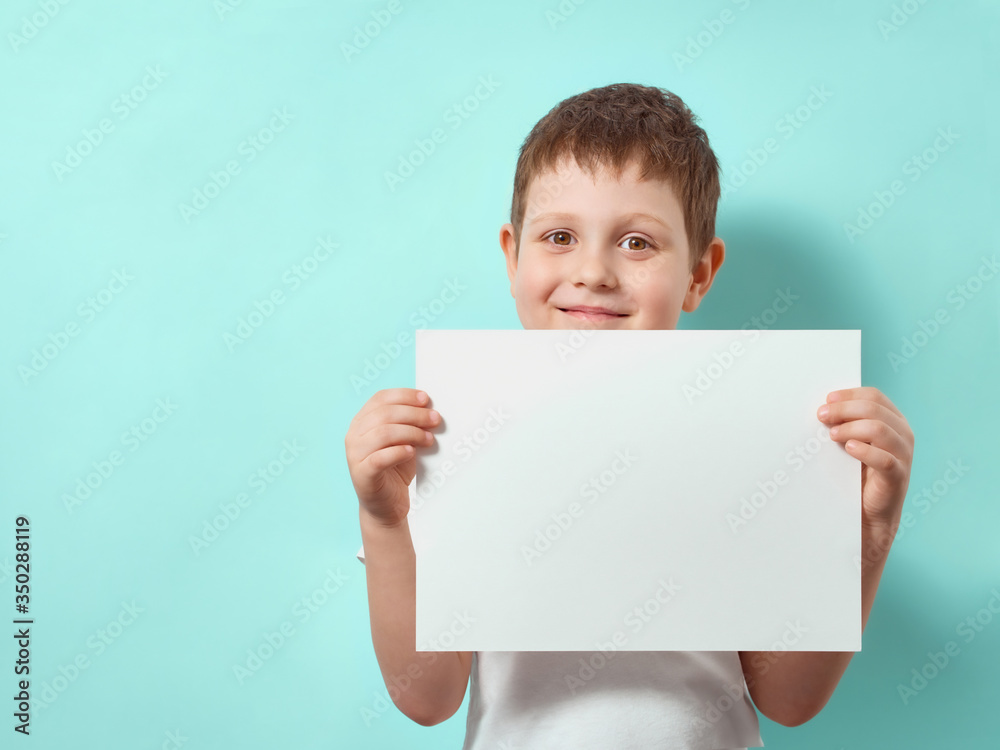 Wall mural four-year-old boy smiles and holds blank white paper sheet. happy child on blue background with copy