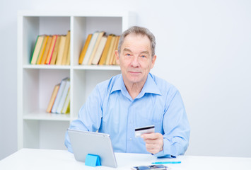 Senior citizen man working on a tablrt holding glasses on his forehead looking at a credit card in his hand