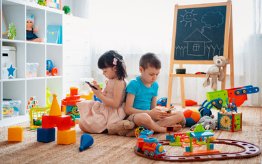 Siblings children brother and sister, friends sit on the floor of the house in the children's play room with smartphones, detached from the scattered toys. - obrazy, fototapety, plakaty
