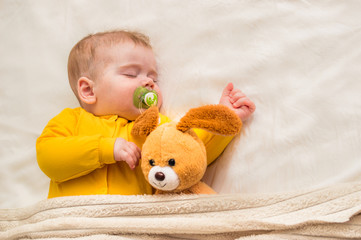 Close-up portrait of a sleeping infant in an embrace with a toy and a pacifier in his mouth.