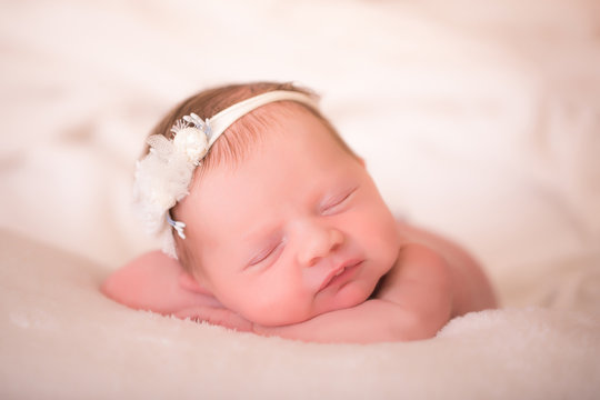 A sleeping two week old newborn baby girl wearing a vintage lace headband with flower. She is wrapped in gauzy cream colored fabric and sleeping on a white billowy blanket.