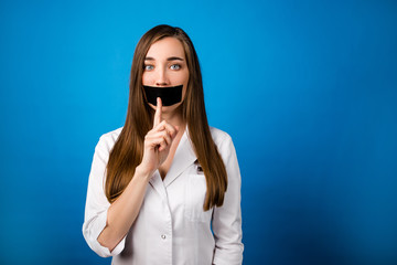 Girl doctor in a white coat with black tape over her mouth on a blue background close-up, silence...
