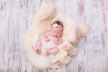 A portrait of a beautiful, seven day old, newborn baby girl wearing a large, fabric rose headband. She is swaddled with gauzy fabric and sleeping in a wire basket.