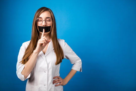 Beautiful girl doctor in a white coat with duct tape over her mouth on a blue background, silence and non-disclosure of medical secrets