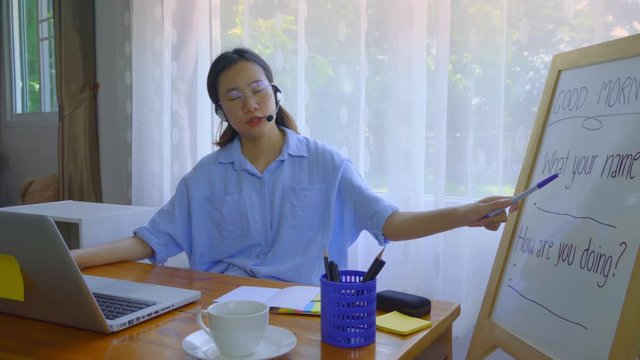 An Asian woman is teaching online via a laptop via webcam.