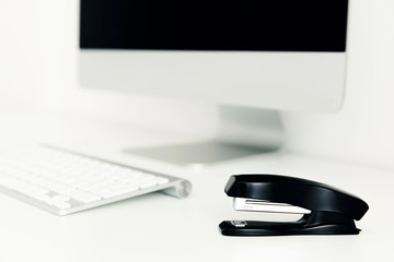Black paper stapler is on white table, against background of monitor and keyboard. Office concept....