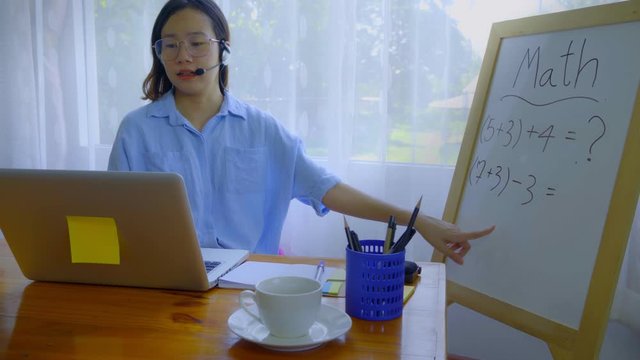An Asian woman is teaching online via a laptop via webcam.