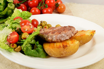 two pieces of grilled potato and grilled meat next to lettuce with tomatoes and olives on a white plate